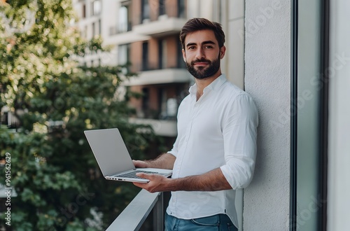 Young businessman holding laptop while standing on balcony in front of modern office building