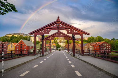 Historic Old Town Bridge or Gamle Bybro Bridge in Trondheim, Norway, crossing the Nidelva River with colorful wooden houses and a rainbow in the background photographed at sunset.