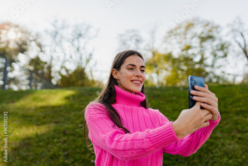 young smiling woman in pink sweater walking in green park using phone