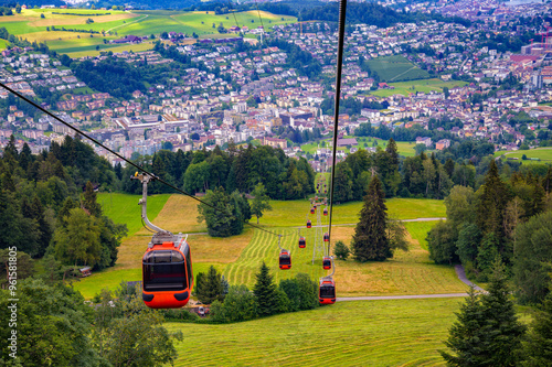 Gondolas ascending to Mount Pilatus, with the city of Kriens near Lucerne in the background, Switzerland, surrounded by lush green fields and forested areas. photo