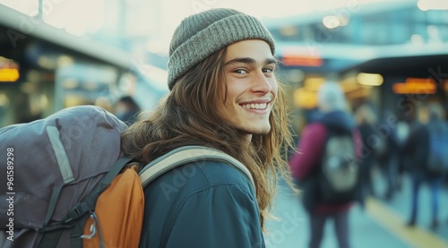 A young traveler with long hair and beanie stands at the train station, smiling as he looks towards his friends waiting for their turn to board the bus. 