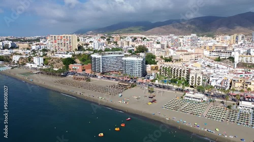 Drone flight along the coastline of Torremolinos over Bajondillo beach with sun loungers on the sand, city scape and mountains in the background.