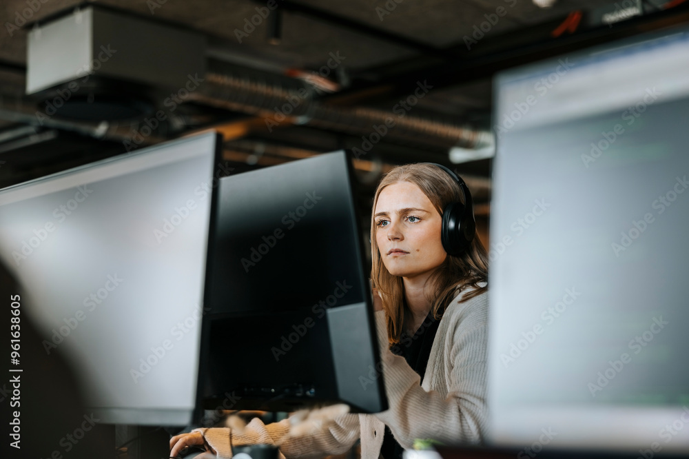 © Maskot - Focused female entrepreneur with wireless headphones doing coding on computer in office