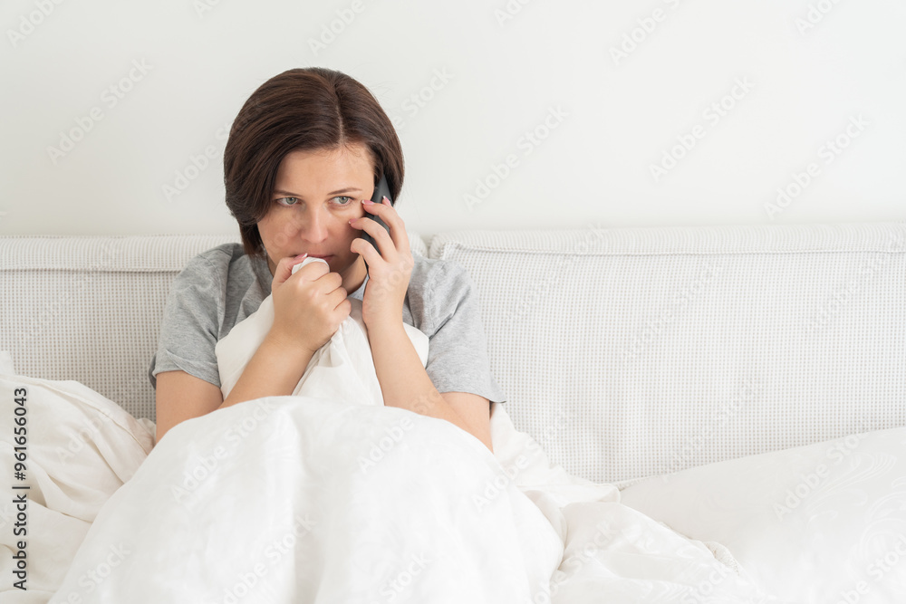 Woman using smartphone as she sits in bed covered with duvet, scared and terrified Woman using smartphone as she sits in bed covered with duvet, scared and terrified