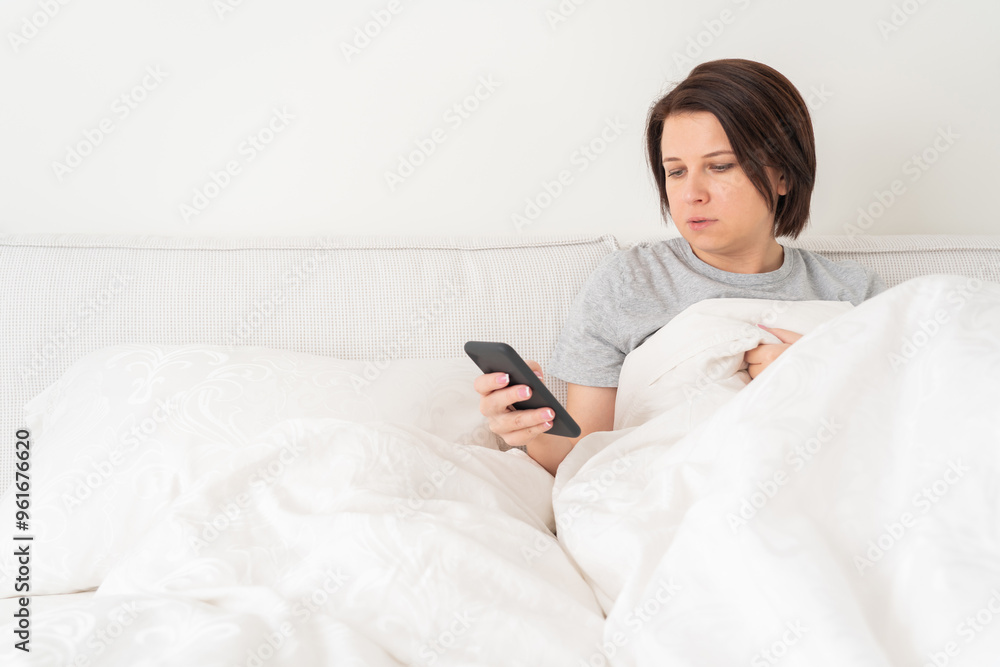 Woman using smartphone as she sits in bed covered with duvet Woman using smartphone as she sits in bed covered with duvet