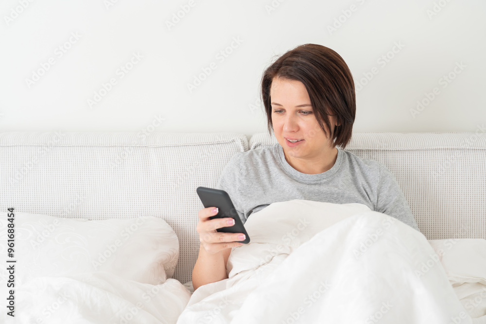 Woman using smartphone as she sits in bed covered with duvet Woman using smartphone as she sits in bed covered with duvet