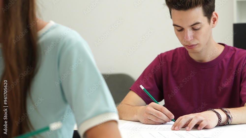 High school teenagers in classroom, boy at desk sitting writing exam checking tasks on paper sheets. Modern education concept. High school teenagers in classroom, boy at desk sitting writing exam checking tasks on paper sheets. Modern education concept.