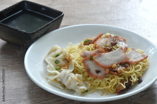 dry Chinese egg yellow noodles topping red barbecue pork and dumpling on plate with clear soup in bowl