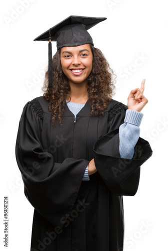 Young hispanic woman wearing graduation uniform very happy pointing with hand and finger to the side