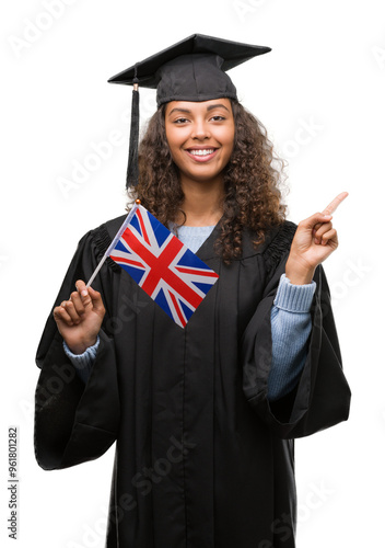 Young hispanic woman wearing graduation uniform holding flag of UK very happy pointing with hand and finger to the side