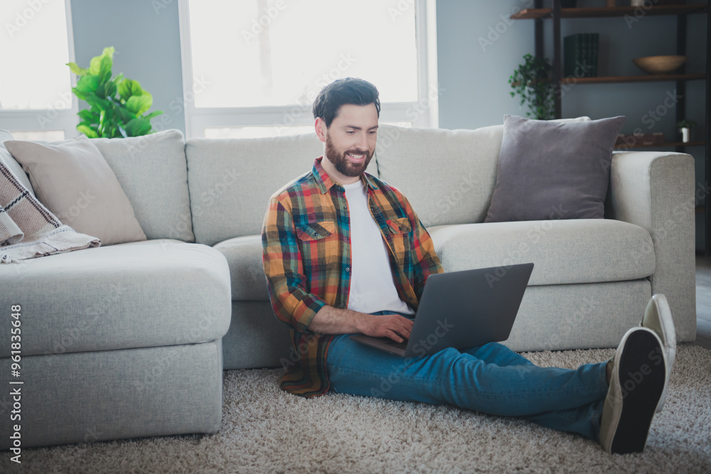 Full length photo of attractive young male sit floor netbook wear checkered shirt cozy day light home interior living room