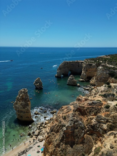 The photo shows the impressive limestone arches near Benagil Cave in the Algarve, Portugal. Crystal-clear green waters flow beneath the towering cliffs, set against a bright blue sky and coastline.