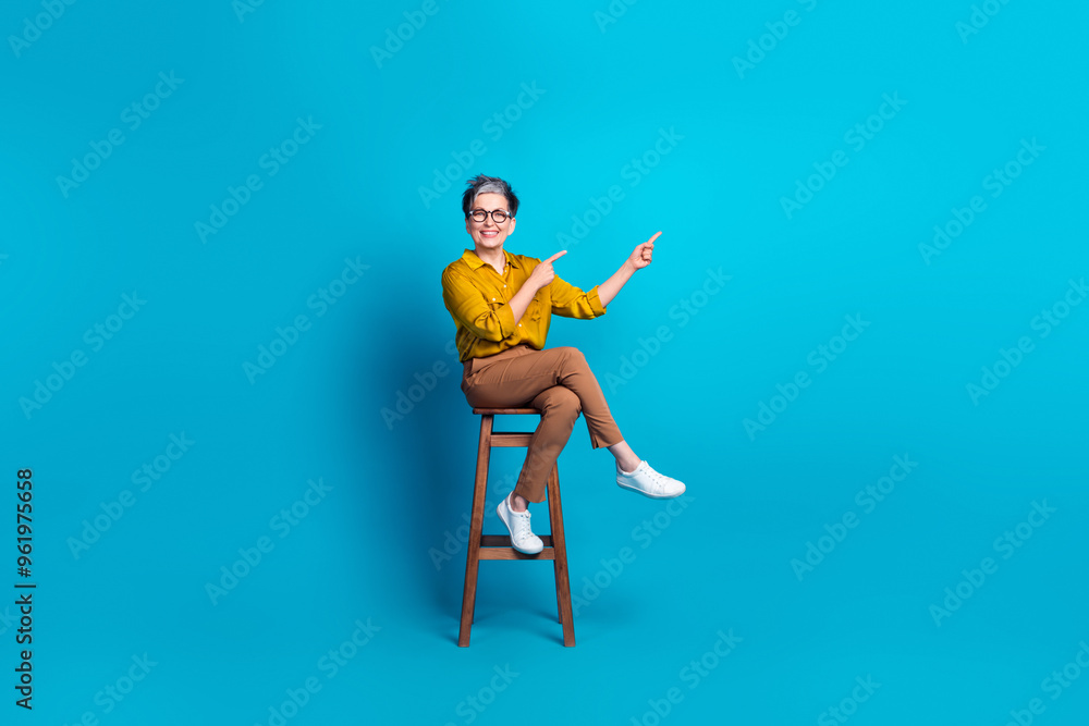 Obraz premium Full length photo of charming positive woman wear blue blouse eyeglasses sitting chair pointing empty space isolated blue color background