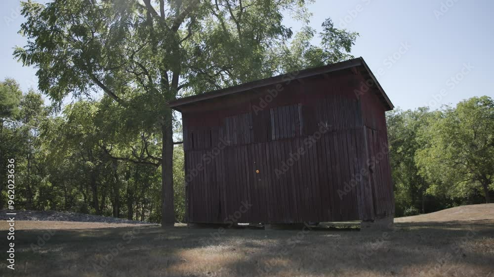 Old wooden barn shed on private property beneath some trees and forested landscape with sun flares in slow motion as the wood paneling and red oak building structure is still and olden Old wooden barn shed on private property beneath some trees and forested landscape with sun flares in slow motion as the wood paneling and red oak building structure is still and olden