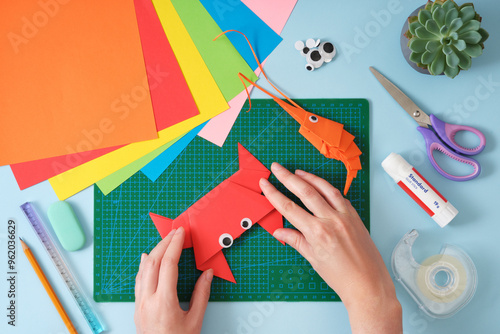 Top view of paper cutting tools, scissors, cutter and paper crafts. Women's hands fixing the final folds for the crab toy. DIY project concept for kids. Flat lay