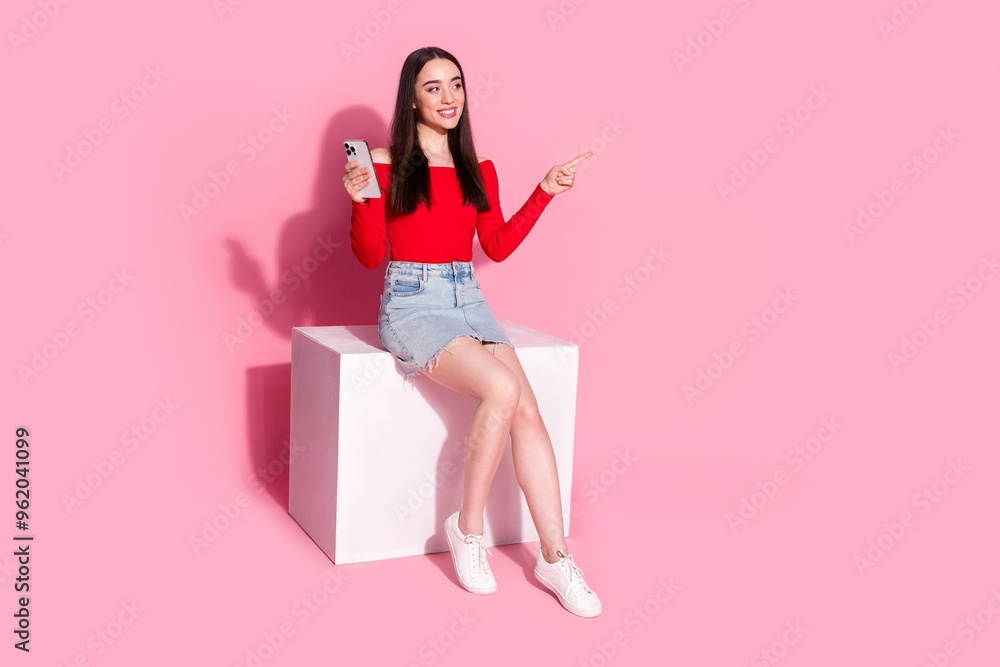 Full body portrait of pretty young girl sit cube hold phone direct finger empty space isolated on pink color background Full body portrait of pretty young girl sit cube hold phone direct finger empty space isolated on pink color background