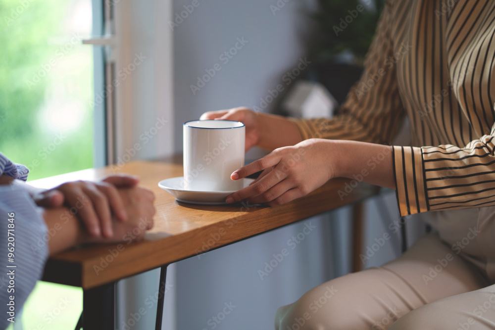 Two people are sitting at a table with a white cup and saucer. One of them is holding the cup and saucer