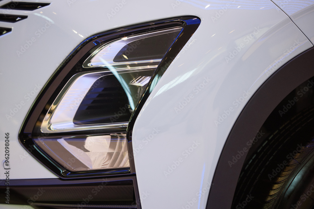 Close up on headlight of modern automobile in futuristic design. White demo vehicle with shiny coat displayed in car showroom at dealership center Close up on headlight of modern automobile in futuristic design. White demo vehicle with shiny coat displayed in car showroom at dealership center