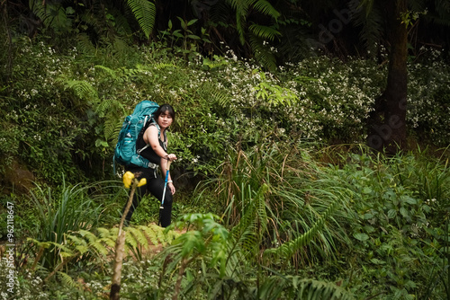 Young asian woman wearing carrier backpack is doing solo hiking activity at Mount Prau, Dieng. She walks along a narrow path with grass and trees beside it. Concept for outdoor activity photo