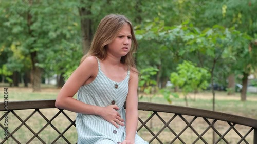 Kid girl sitting alone on the bench in the park, in pain holding belly, feeling stomach ache, symptoms or diarrhea. Upset preteen girl suffering from indigestion, flatulence and abdominal problem