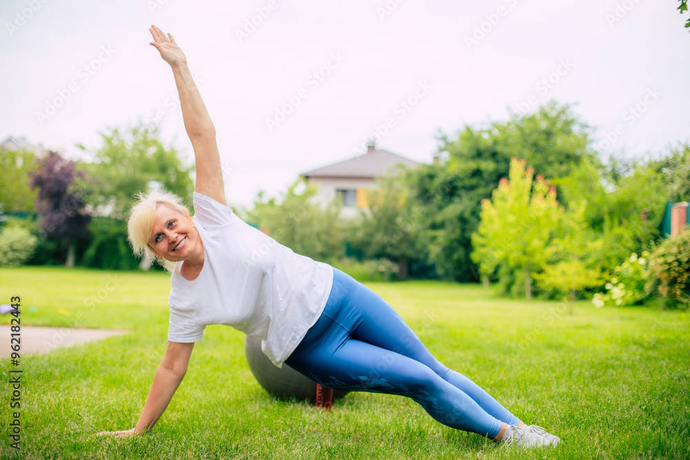Adult woman practices a side plank exercise outdoors on a grassy lawn, smiling as she holds her balance. The image captures a moment of fitness and well-being in a serene natural setting. Adult woman practices a side plank exercise outdoors on a grassy lawn, smiling as she holds her balance. The image captures a moment of fitness and well-being in a serene natural setting.