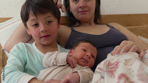 Older sibling gently cradles baby while resting on mother’s lap. The siblings share a peaceful moment, highlighting the close bond and affection between them in a cozy home environment