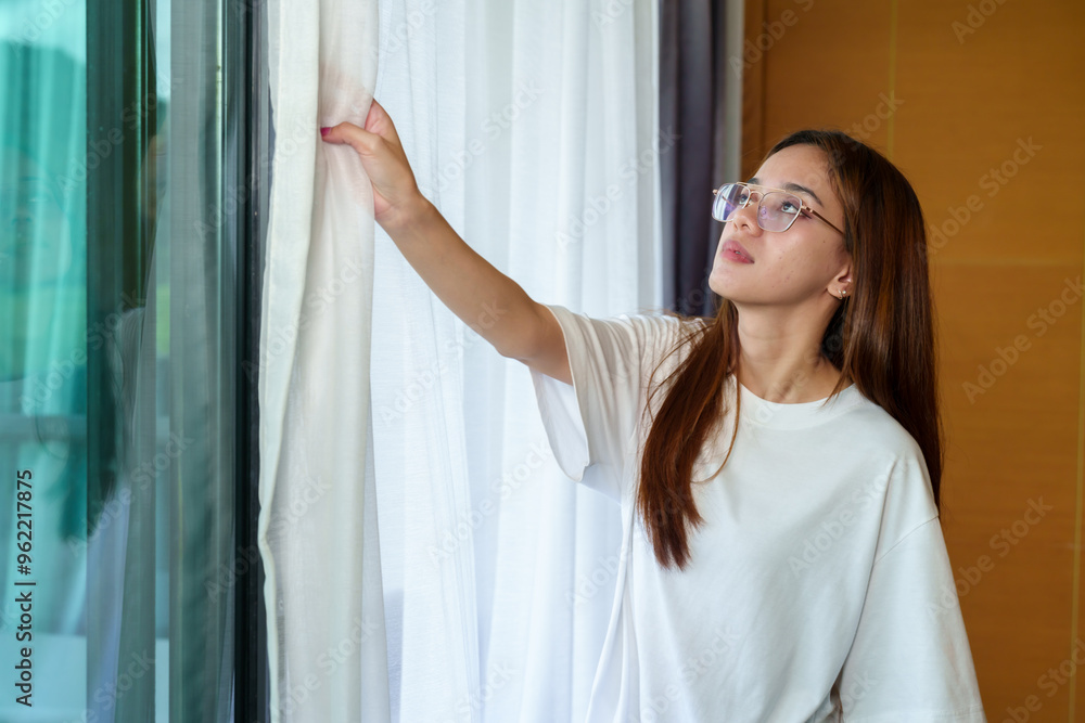 Young Asian woman opening bedroom curtains to let in natural light. standing by the window, adjusting the curtains with a focused expression. Soft daylight entering the room, home setting Young Asian woman opening bedroom curtains to let in natural light. standing by the window, adjusting the curtains with a focused expression. Soft daylight entering the room, home setting