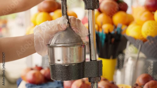 Street vendor wearing gloves prepares fresh pomegranate juice using manual fruit press, showcasing traditional method of extracting juice from ripe pomegranates in bustling market photo