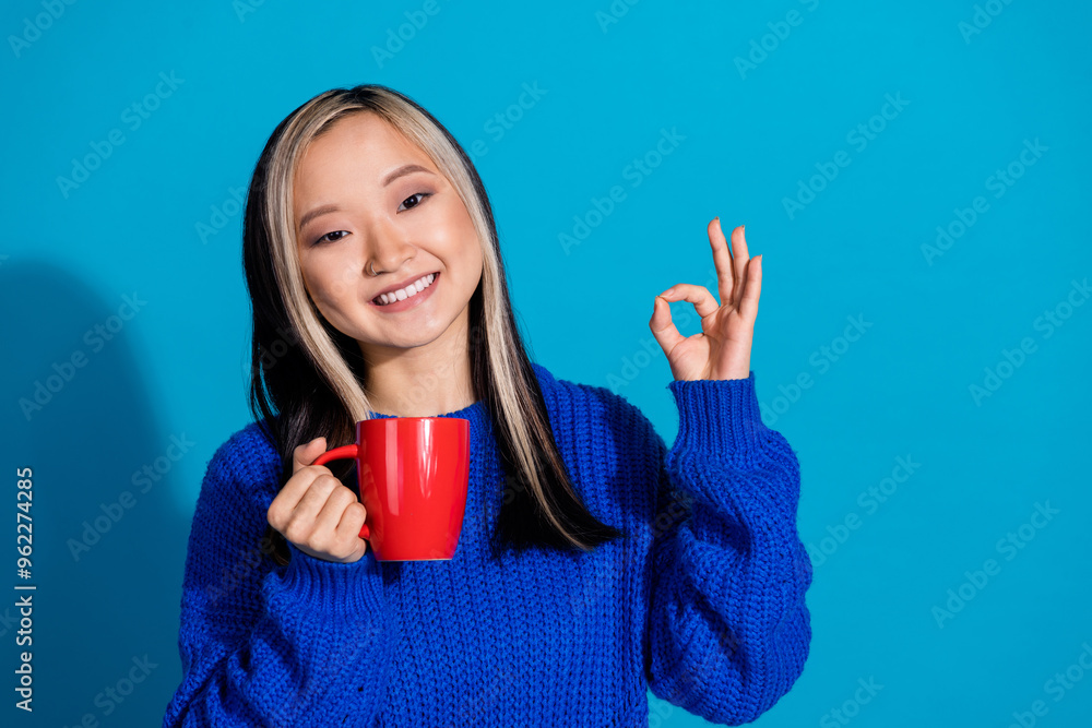 Photo of cute thai girl wearing knitted warm sweater showing okey sign likes cappuccino coffee isolated on blue color background