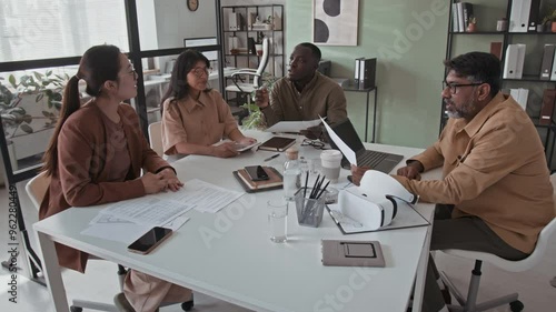 Full shot of multiethnic businesspeople discussing new project or startup sitting at table with documents, laptop and vr headset in modern office photo