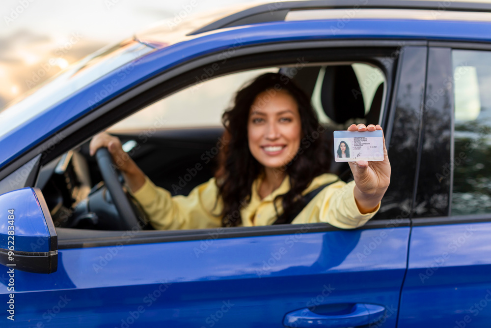 Cheerful young lady sitting in blue car and showing her new driving license, selective focus. Woman pass driving test, driver courses, exam concept Cheerful young lady sitting in blue car and showing her new driving license, selective focus. Woman pass driving test, driver courses, exam concept