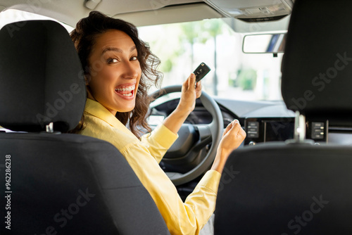 Excited European woman sitting in car salon, happy owner holding car key looking back at camera and smiling. Car insurance concept photo