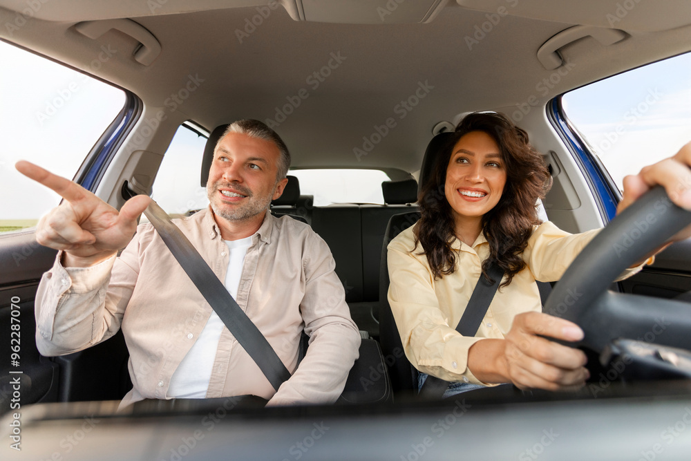 Happy middle aged man pointing with finger at the road and saying something while woman driving car, enjoying trip together