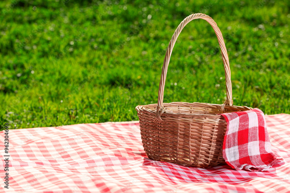 One picnic wicker basket with checkered napkin and blanket on green grass. Space for text One picnic wicker basket with checkered napkin and blanket on green grass. Space for text