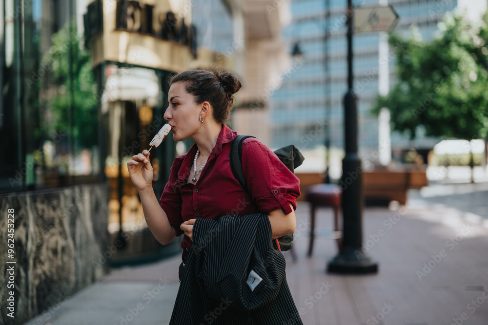 Fototapeta premium Young businesswoman savoring ice cream while walking in a city setting, encapsulating a blend of professionalism and casual leisure.