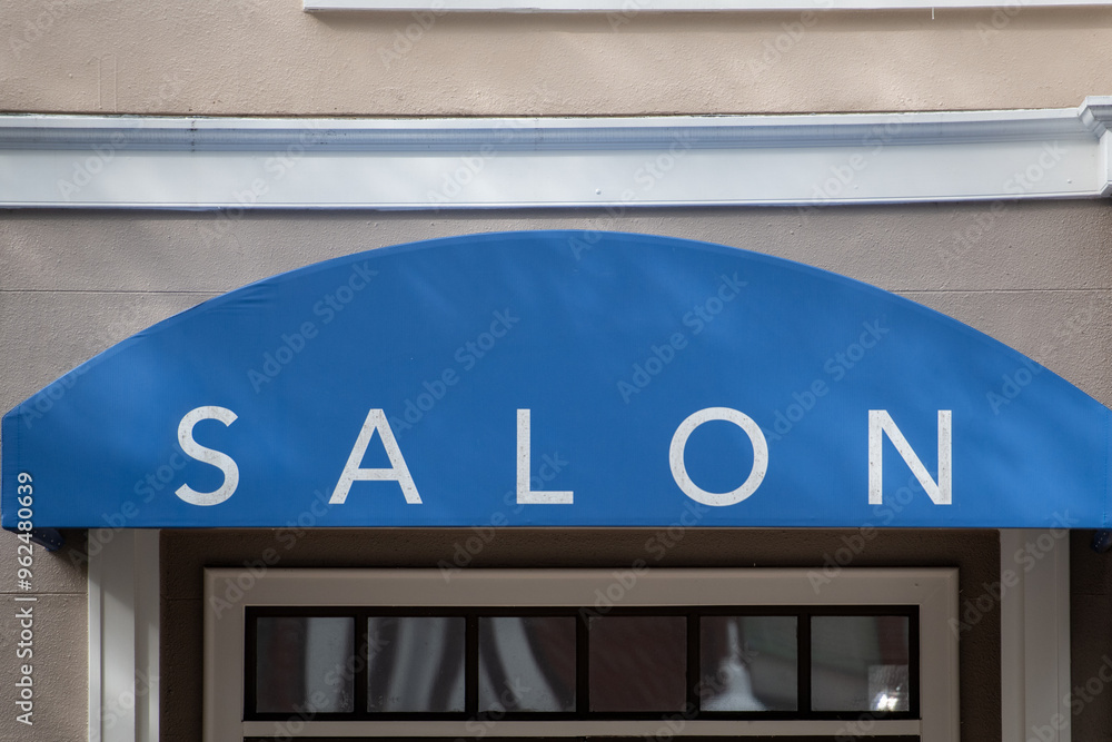 A blue cloth awning with salon in white letters over a metal framed glass door of a business. The exterior wall is beige with white trim. The sign is an arched storefront commercial canopy. A blue cloth awning with salon in white letters over a metal framed glass door of a business. The exterior wall is beige with white trim. The sign is an arched storefront commercial canopy.