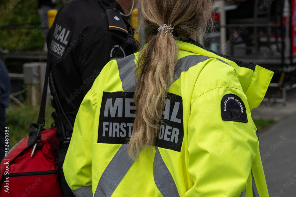 Close-up of a medical first responder wearing a bright yellow coat with a grey reflective cross. The person is back on to the camera. The young female front line worker has a long blond ponytail. Close-up of a medical first responder wearing a bright yellow coat with a grey reflective cross. The person is back on to the camera. The young female front line worker has a long blond ponytail.