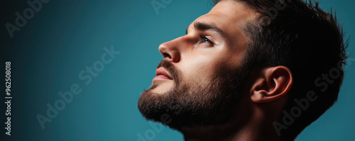 Close-up side profile of a young man with a beard gazing upwards against a blue background, showcasing contemplation.