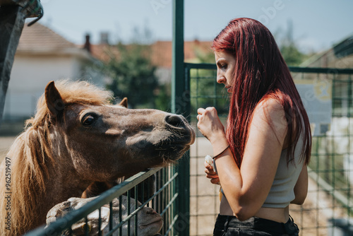 A young woman interacts with a pony at a petting zoo, feeding it by hand. The sunny outdoor setting evokes a sense of calm and peacefulness. photo