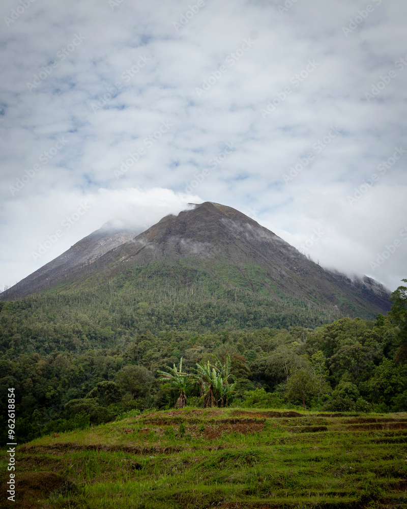 Mount Sinabung, an active volcano in North Sumatra, Indonesia, with smoke rising from its summit, surrounded by lush greenery and rugged volcanic rocks under a dramatic, cloud-filled sky. Mount Sinabung, an active volcano in North Sumatra, Indonesia, with smoke rising from its summit, surrounded by lush greenery and rugged volcanic rocks under a dramatic, cloud-filled sky.