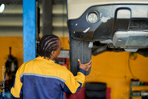 African american female in technicial uniform checking customer car tire whil car on machine power lift in car garage. photo