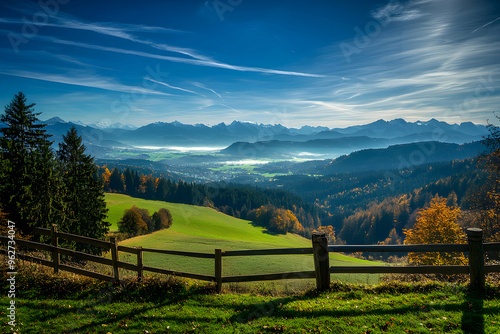 A picturesque view of a rolling green hillside with a wooden fence in the foreground and a valley surrounded by mountains in the distance.