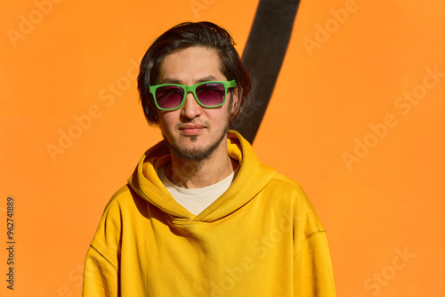 Stylish Asian guy in green glasses stands against a yellow wall,