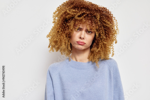 Sad young woman with curly hair in a light blue sweater, looking directly at the camera against a neutral background Her expression conveys a sense of disappointment or introspection