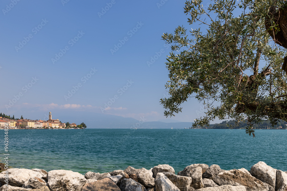 Picturesque Salo on Lake Garda viewed through an olive branch, with a rocky foreground and distant mountains under a clear sky Picturesque Salo on Lake Garda viewed through an olive branch, with a rocky foreground and distant mountains under a clear sky