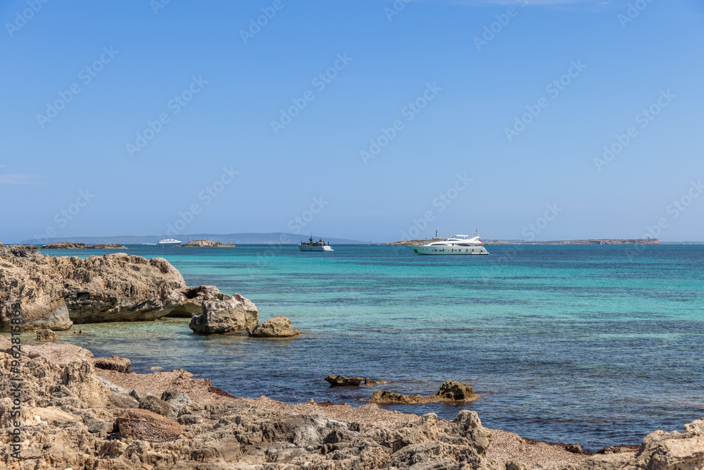 The rocky coastline of Ibiza with yachts anchored in the clear turquoise waters, reflecting a luxurious and tranquil vacation atmosphere The rocky coastline of Ibiza with yachts anchored in the clear turquoise waters, reflecting a luxurious and tranquil vacation atmosphere