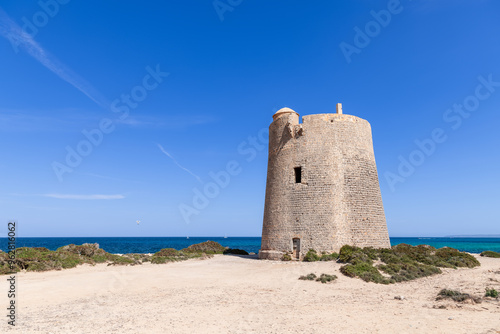 The Torre de ses Portes stands prominently on the sandy coastline of Ibiza, Spain. The stone tower is framed by a clear blue sky and the sparkling Mediterranean Sea, creating a serene coastal scene photo