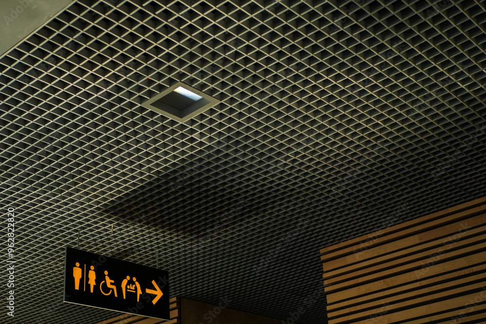 A toilet sign on a ceiling with a person and a wheelchair symbol and a sign with an arrow pointing to the right in a public office or airport. Travel and tourism A toilet sign on a ceiling with a person and a wheelchair symbol and a sign with an arrow pointing to the right in a public office or airport. Travel and tourism