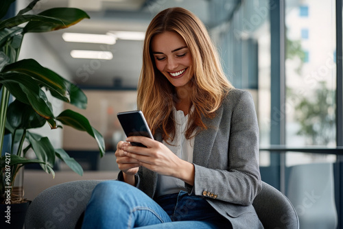 A smiling young woman in a grey blazer sits in a modern office and looks at her smartphone.