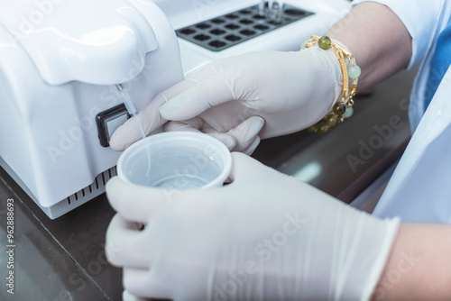 A person wearing gloves holds a cup of distilled water under a semi-auto chemistry analyzer. Aspirating water with a hose. At a medical laboratory or hospital. photo